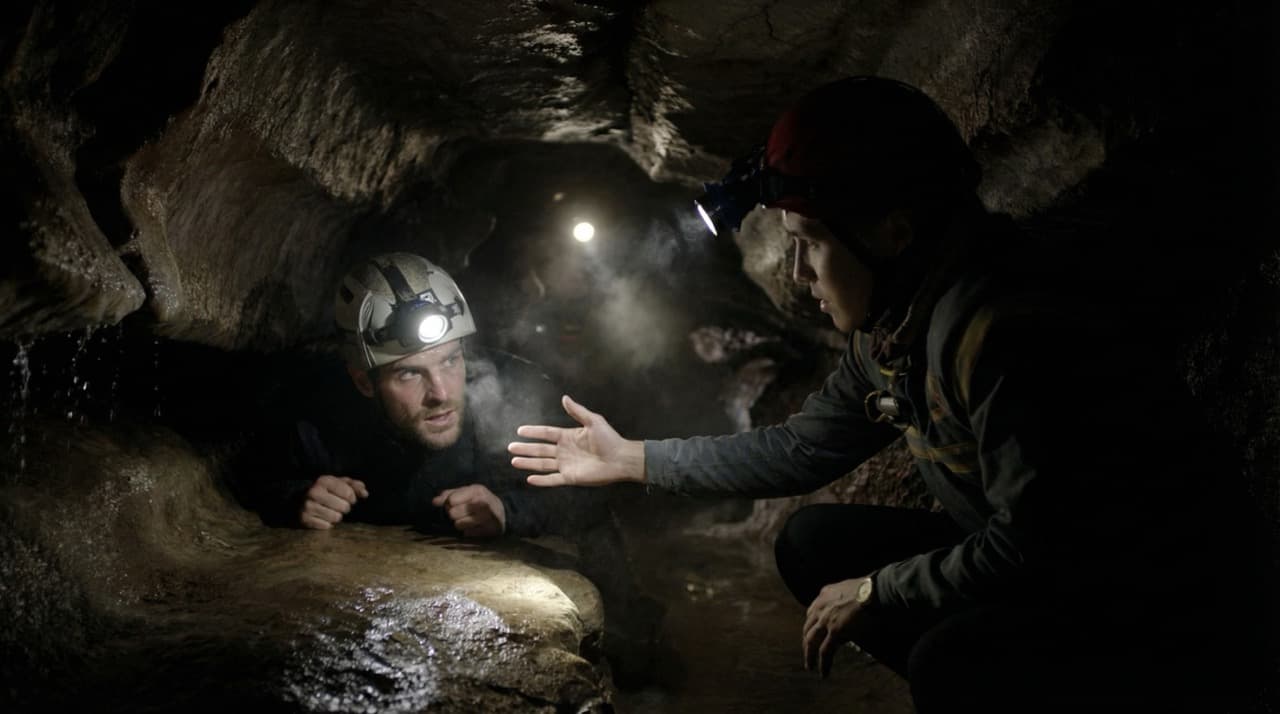 Instructor guiding a participant through a tight cave passage — headlamps, wet limestone, mutual trust